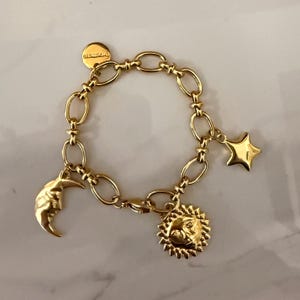 Könnte beinhalten: Ein goldfarbenes Gliederkettarmband mit drei verschiedenen Anhängern: einem Halbmond mit Gesicht, einer Sonne mit Gesicht und einem fünfzackigen Stern. Ein kleiner runder Anhänger mit der Aufschrift "GLAMIRA" ist am Verschluss befestigt.