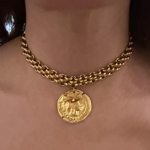 Puede incluir: Un collar tipo gargantilla de tono dorado con un diseño de cadena gruesa de múltiples capas. Un gran colgante circular de tono dorado con un retrato de perfil en relieve y detalles intrincados cuelga del centro de la cadena sobre el cuello.