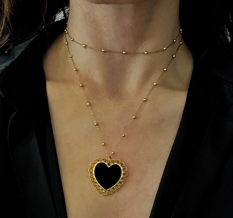 Pu&ograve; includere: Una collana con catena dorata con un ciondolo a forma di cuore nero. Il ciondolo ha un design delicato e decorato.