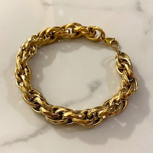 Op de afbeelding: Een goudkleurige kettingarmband met dikke, in elkaar grijpende ovale schakels met een gefacetteerd, getextureerd oppervlak. De armband is gemaakt van gepolijst metaal en bevat een veilige karabijnsluiting aan één uiteinde om het sieraad te sluiten.