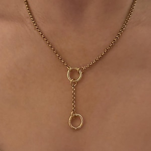 May include: A gold-toned lariat necklace featuring a delicate rolo chain. The design includes a central circular ring connector that joins the main chain to a shorter vertical drop chain, which terminates in a smaller circular pendant set with tiny clear stones.