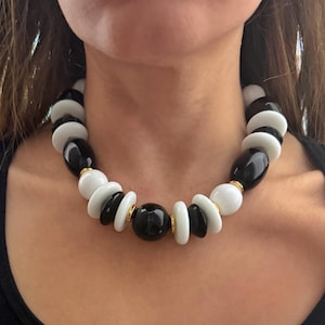 May include: A statement necklace with a bold design. It features a mix of black and white beads in various shapes, including round and oval. Gold-toned spacers add detail. The necklace is worn around the neck.