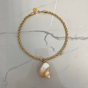 May include: A gold rope chain necklace with a large seashell pendant. The seashell is white with a pink hue and has a spiral pattern.