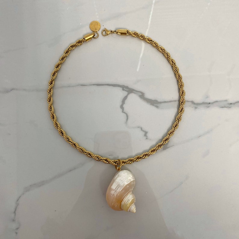May include: A gold rope chain necklace with a large seashell pendant. The seashell is white with a pink hue and has a spiral pattern.