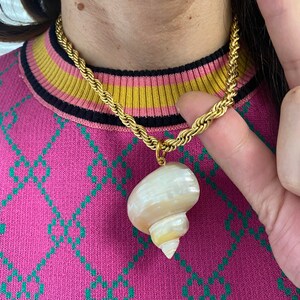 May include: A gold chain necklace with a large, white seashell pendant. The necklace is worn by a person with a pink sweater.