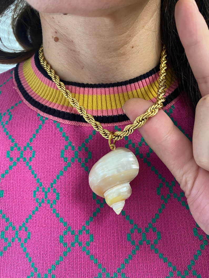 May include: A gold chain necklace with a large, white seashell pendant. The necklace is worn by a person with a pink sweater.