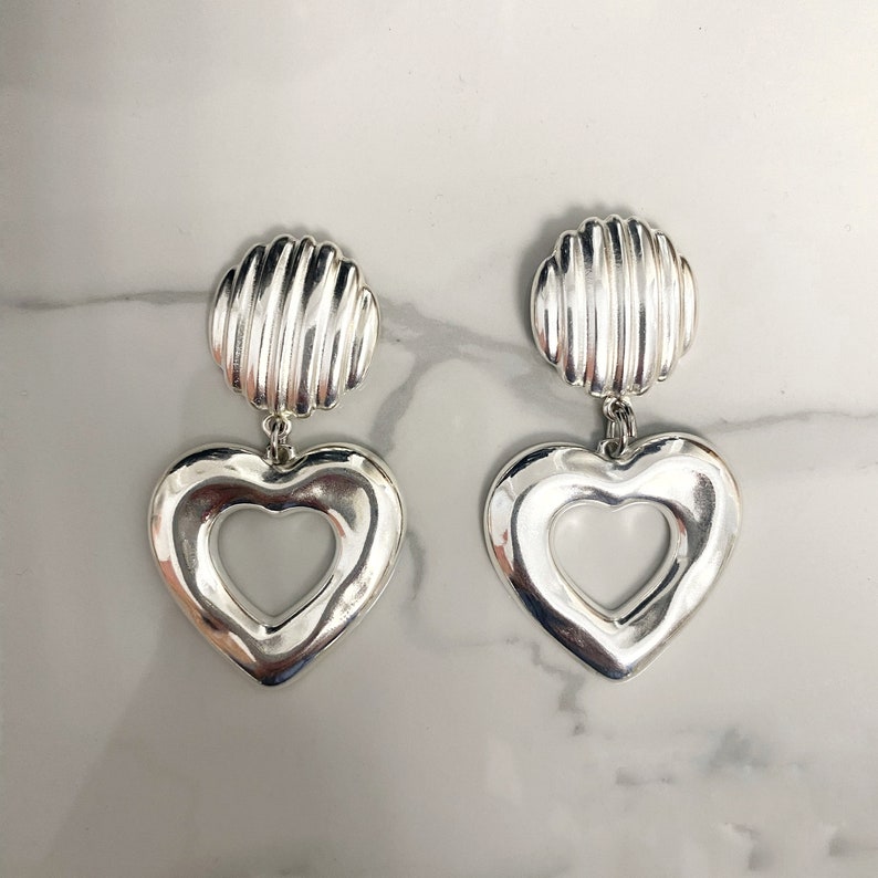 May include: A pair of silver heart-shaped earrings with a textured round disc at the top.