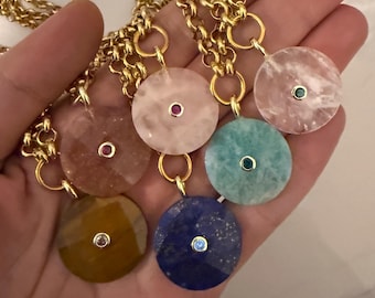 May include: A collection of gold chain necklaces, each featuring a large, round pendant in various colors: pink, blue, green, and yellow. Each pendant has a small, contrasting gemstone in the center. The necklaces are displayed in a hand.