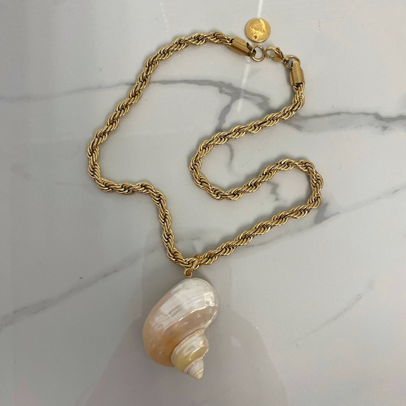 May include: A gold chain necklace with a large seashell pendant. The chain is a rope style and the seashell is a light pink and white color.