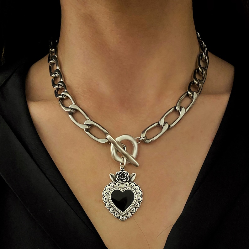 May include: A silver chain necklace with a large, chunky link design. The necklace has a toggle clasp and a pendant in the shape of a black heart with a silver rose detail.