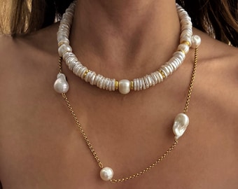 May include: A two-strand necklace featuring white, disc-shaped beads and large, irregular pearls. The necklace has gold-colored accents and a delicate gold chain. The jewelry is worn around the neck.