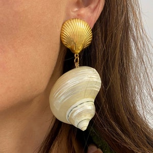 Op de afbeelding: Gouden en witte schelp oorbellen. De bovenkant van de oorbel is een gouden schelp met een getextureerd oppervlak. De onderkant van de oorbel is een witte schelp met een spiraalvormig patroon.