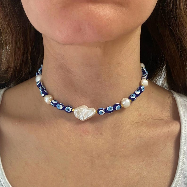 K&ouml;nnte beinhalten: Eine blaue und wei&szlig;e Perlenkette mit einem gro&szlig;en wei&szlig;en Perlenanh&auml;nger. Die Perlen haben die Form von b&ouml;sen Augen.
