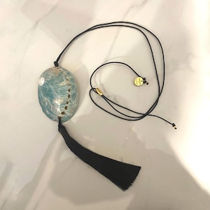 Könnte beinhalten: Abalone-Muschel-Anhänger-Halskette mit schwarzer Quaste und Kordel. Die Muschel hat einen türkisblauen und schillernden Glanz. Ein kleiner goldfarbener Anhänger ist an der Kordel befestigt.