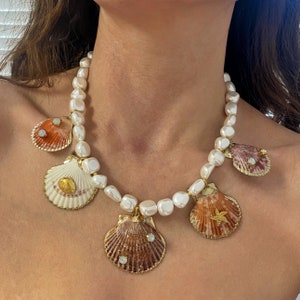 Puede incluir: Un collar de perlas blancas con cinco colgantes de concha dorada. Las conchas están decoradas con diamantes de imitación y una estrella de mar.