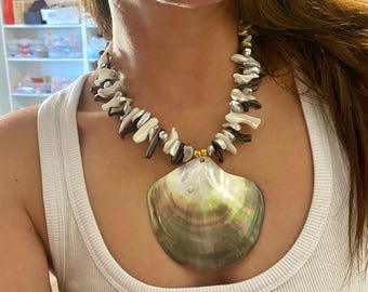 May include: A statement necklace featuring a large, iridescent seashell pendant. The necklace has a strand of white and dark brown shell fragments. The pendant is a mix of green, gray, and white. The necklace is worn on a white tank top.