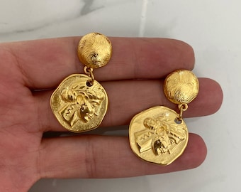 May include: Gold-toned earrings featuring a circular top with a textured swirl design and a dangling coin-shaped pendant. The pendant displays a detailed relief of a classical figure with a laurel wreath. The earrings have a vintage aesthetic.