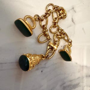 Könnte beinhalten: Ein goldfarbenes Kettenarmband mit drei hängenden Anhängern. Zwei Anhänger sind flache, rechteckige grüne Steine mit strukturierten goldenen Schlaufen, und ein Anhänger ist ein runder grüner Stein in einer verzierten, strukturierten Goldfassung.