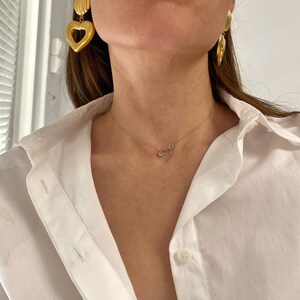 May include: A woman wearing a white button-down shirt with a gold heart-shaped pendant necklace and gold heart-shaped earrings.