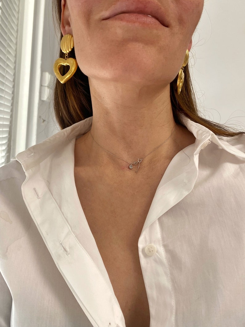 May include: A woman wearing a white button-down shirt with a gold heart-shaped pendant necklace and gold heart-shaped earrings.
