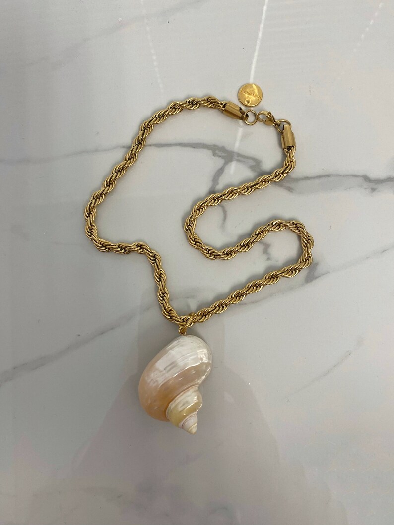 May include: A gold chain necklace with a large seashell pendant. The seashell is white with pink and yellow stripes.