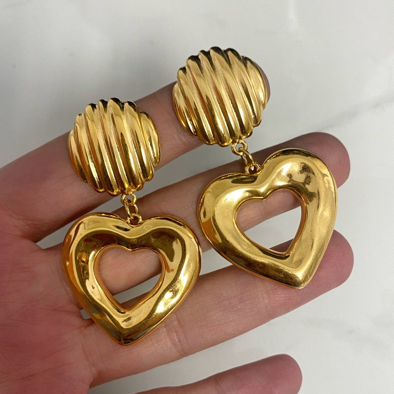 May include: A pair of gold-tone earrings with a heart-shaped design. The hearts have a cutout in the center, creating a second heart shape. The earrings are attached to a round, ribbed disc.