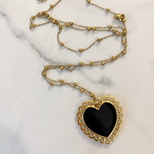 Pu&ograve; includere: Una collana a catena dorata con un grande ciondolo a forma di cuore nero. Il ciondolo a forma di cuore ha un design delicato e decorato attorno al bordo.