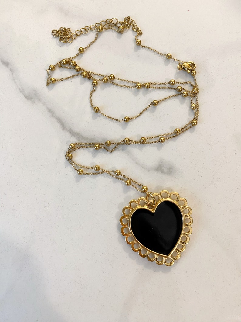 Pu&ograve; includere: Una collana a catena dorata con un grande ciondolo a forma di cuore nero. Il ciondolo a forma di cuore ha un design delicato e decorato attorno al bordo.