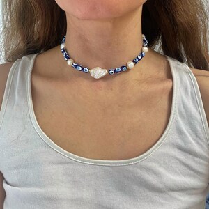 K&ouml;nnte beinhalten: Eine blaue und wei&szlig;e Perlenkette mit einem gro&szlig;en wei&szlig;en Perlenanh&auml;nger. Die Kette hat ein sich wiederholendes Muster aus blauen b&ouml;sen Augen.
