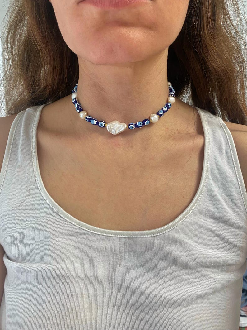 K&ouml;nnte beinhalten: Eine blaue und wei&szlig;e Perlenkette mit einem gro&szlig;en wei&szlig;en Perlenanh&auml;nger. Die Kette hat ein sich wiederholendes Muster aus blauen b&ouml;sen Augen.