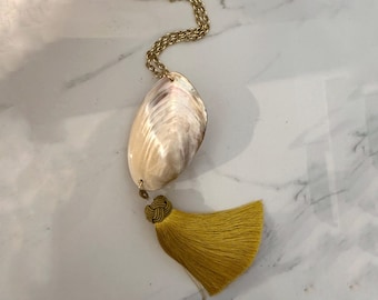 May include: A necklace featuring a gold-toned chain, a large iridescent seashell pendant, and a mustard yellow fabric tassel. The tassel is attached to the base of the shell with a decorative knot, creating a long, layered accessory with natural textures.