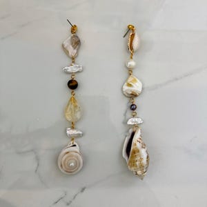 Könnte beinhalten: Asymmetrische Ohrringe mit natürlichen Elementen. Jeder Ohrring hat einen goldfarbenen Stecker mit Perlen, Muscheln und Edelsteinen in Weiß, Braun und Gelb. Die Ohrringe haben ein einzigartiges, organisches Design.
