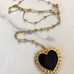 Pu&ograve; includere: Una collana a catena dorata con un grande ciondolo a forma di cuore nero. Il cuore ha un bordo decorativo a forma di conchiglia.