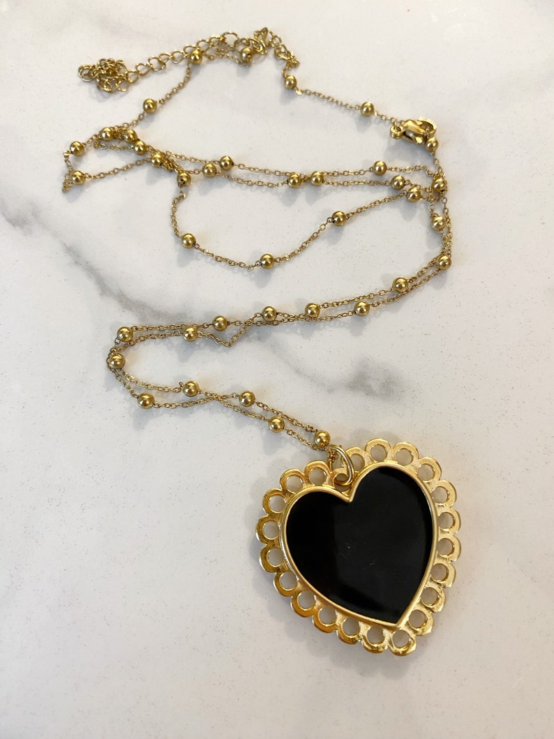 Pu&ograve; includere: Una collana a catena dorata con un grande ciondolo a forma di cuore nero. Il cuore ha un bordo decorativo a forma di conchiglia.