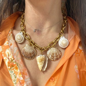 Puede incluir: Un collar de cadena dorada con múltiples dijes de conchas marinas. Los dijes son de varias formas y tamaños, incluyendo un cono, una vieira y una concha en espiral. El collar está en un modelo que lleva una camisa con estampado floral.