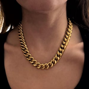 Puede incluir: Un collar de cadena dorado con un diseño de eslabones de cadena gruesos. El collar se lleva alrededor del cuello y descansa sobre una parte superior negra. Los eslabones son gruesos y están estrechamente conectados, creando una pieza audaz.