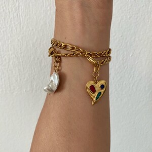 May include: A gold chain bracelet with two charms: a white pearl and a heart-shaped charm with red, blue, and green enamel.
