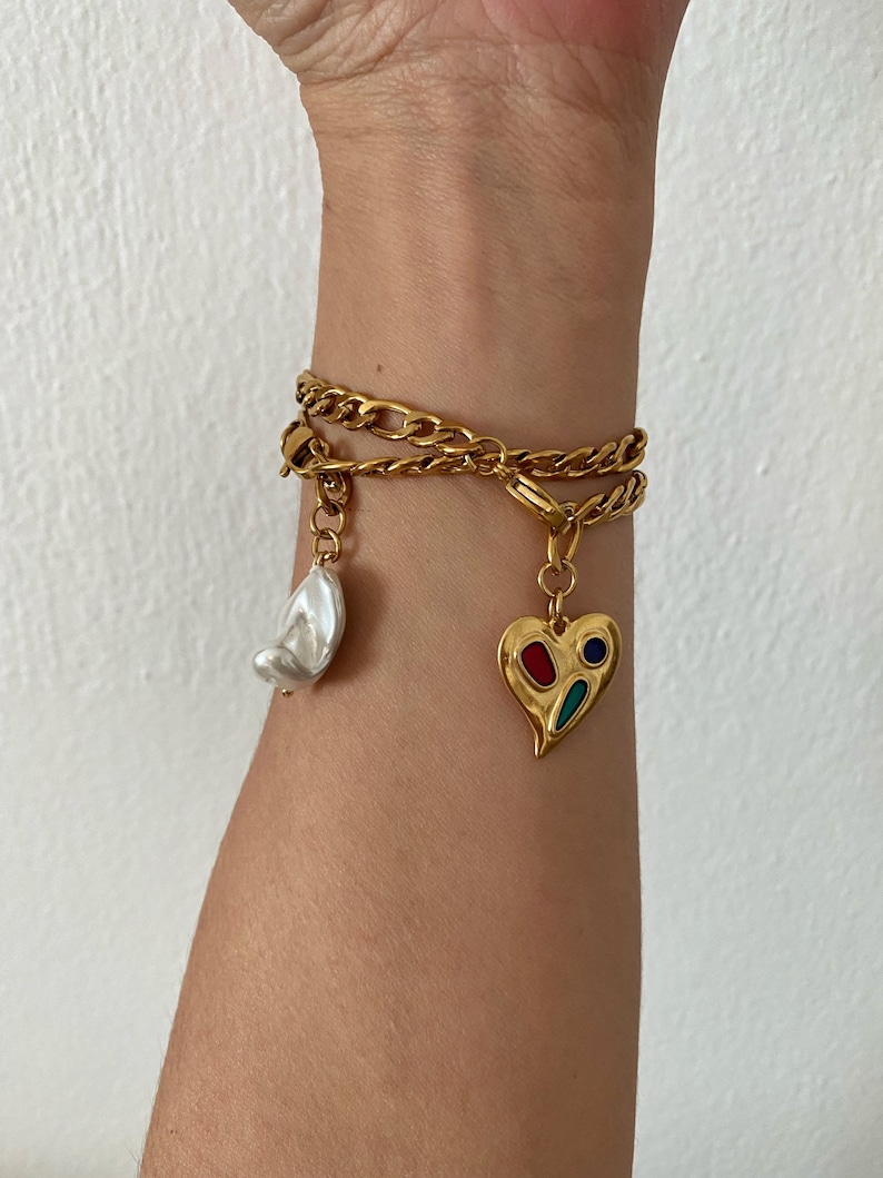 May include: A gold chain bracelet with two charms: a white pearl and a heart-shaped charm with red, blue, and green enamel.