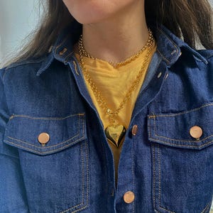 May include: A gold chain necklace with a large heart pendant. The necklace is layered over a yellow t-shirt and a denim jacket.