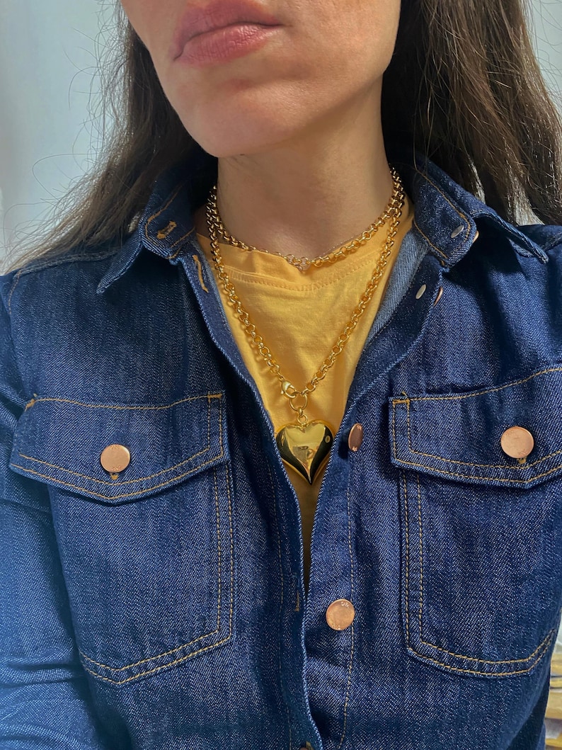 May include: A gold chain necklace with a large heart pendant. The necklace is layered over a yellow t-shirt and a denim jacket.