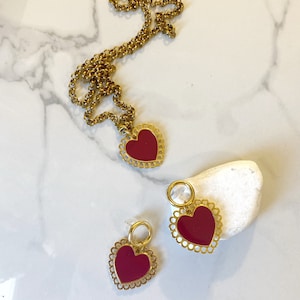 May include: A gold chain necklace with a red heart pendant and a pair of matching red heart earrings. The heart pendants have a delicate, scalloped edge.