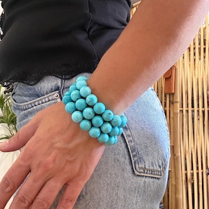 Könnte beinhalten: Ein mehrreihiges Armband bestehend aus drei Reihen runder, leuchtend türkisblauer Perlen mit natürlicher dunkler Maserung. Die Perlen sind einheitlich groß und aneinandergereiht, wodurch ein gestapeltes Erscheinungsbild am Handgelenk entsteht.