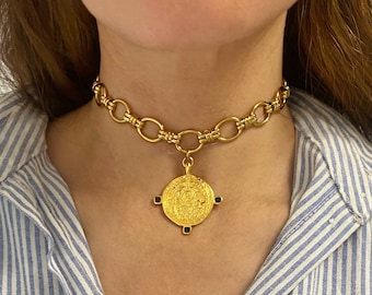 Puede incluir: Un collar gargantilla de cadena dorada con un gran colgante redondo de oro con un diseño intrincado. El colgante tiene cuatro acentos negros.