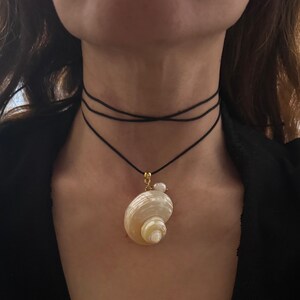 May include: A black cord choker necklace featuring a gold-toned bail attached to a spiral-shaped, iridescent cream-colored seashell pendant. A small, round white pearl is positioned next to the shell, suspended from the thin, multi-layered black cord.