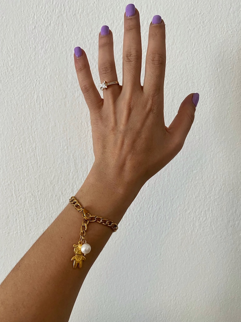 May include: A gold chain bracelet with a small gold bear charm and a white pearl. The bracelet is on a person's wrist.