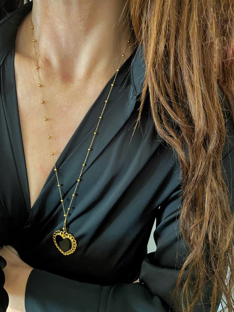 Pu&ograve; includere: Una collana a catena dorata con un ciondolo a forma di cuore nero. Il ciondolo ha un bordo dorato ed &egrave; su uno sfondo nero.
