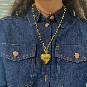 May include: A gold chain necklace with a large, chunky heart pendant. The necklace is layered over a thinner gold chain.