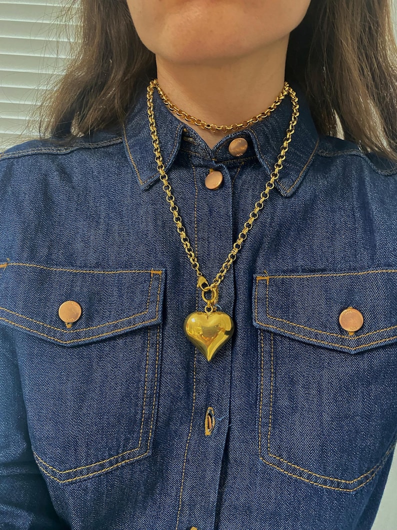 May include: A gold chain necklace with a large, chunky heart pendant. The necklace is layered over a thinner gold chain.