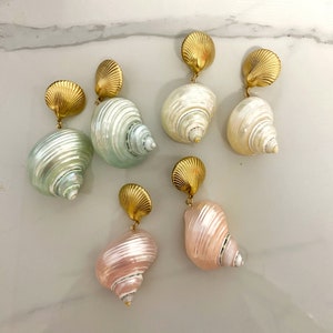 Op de afbeelding: Zes paar gouden oorbellen met schelpvormige bedels. De bedels zijn in tinten wit, roze en groen.