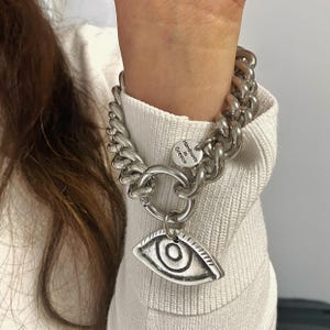 May include: Silver chain bracelet with a large eye charm. The bracelet is made of thick, chunky links and has a tag that says "Handmade in Greece".